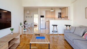 A modern living room with a flat-screen TV, a wooden coffee table, a grey sofa, and a kitchen in the background. - Athens Heartbeat (Athens)