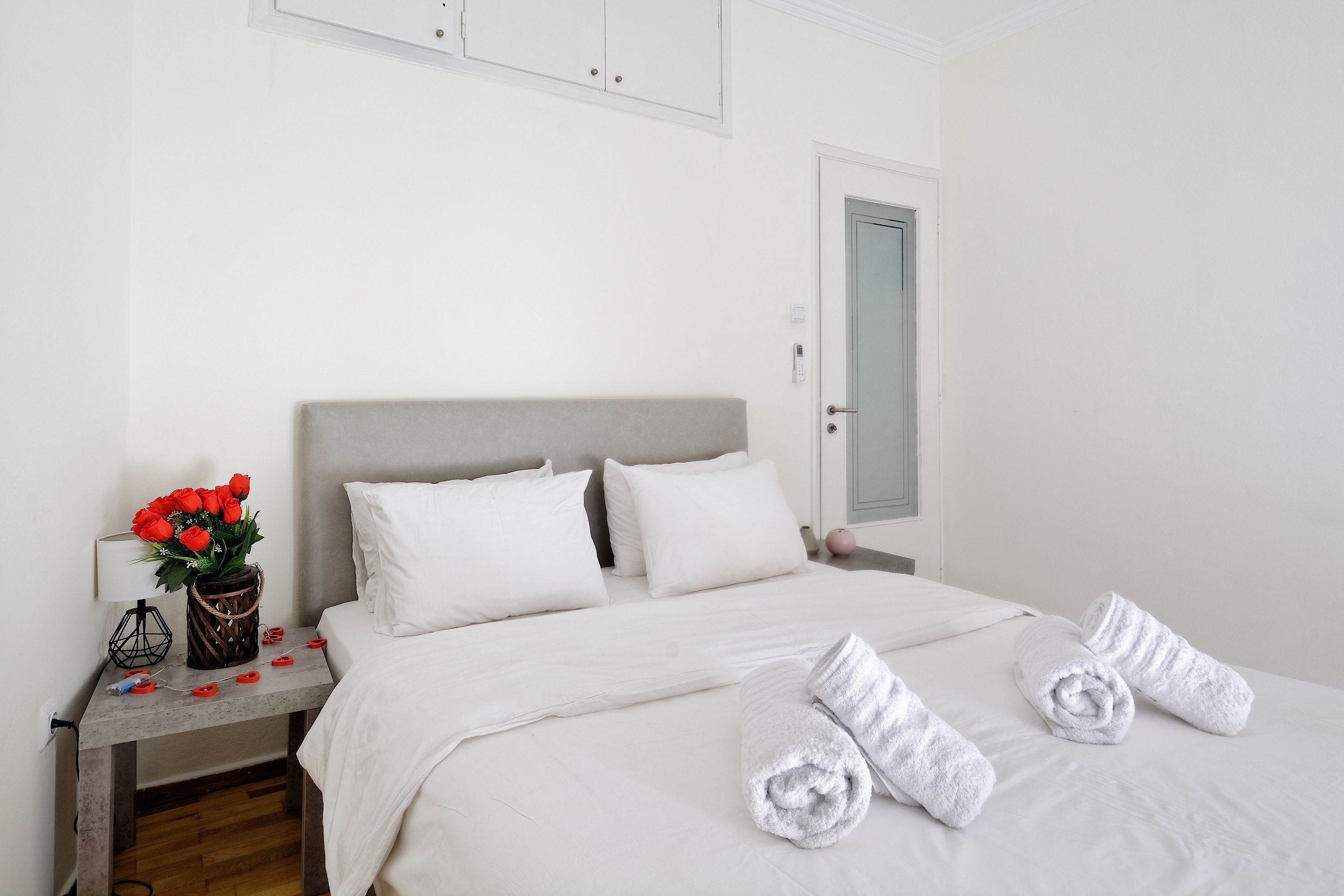 A neatly made bed with white linens, a bedside table with a vase of red flowers, and rolled towels on the bed.