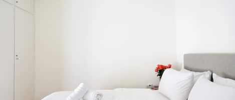 A neatly made bed with white linens and pillows, a grey headboard, and a small bedside table with a vase of red flowers.
