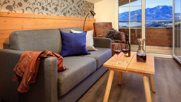 Un salon moderne avec un canapé gris, une tête de lit en bois et une table basse en bois sur laquelle se trouvent des verres à vin et une bouteille.