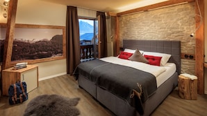 Une chambre avec un grand lit, un mur en pierre, une tête de lit en bois, une vue sur les montagnes et un petit tabouret en bois.