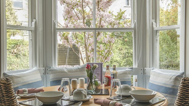 A corner dining area with a round table, wicker chairs, and a view of cherry blossoms through large windows. - Brookdale B&B (Wadebridge)