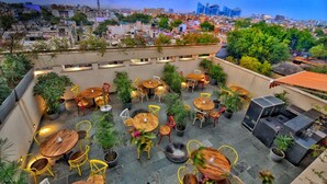 Una terraza en la azotea con varias mesas y sillas redondas, rodeada de plantas en macetas.