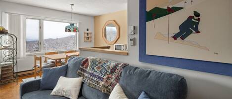 Een gezellige woonkamer met een blauwe bank, een gebreid plaid en uitzicht op een besneeerd landschap door een groot raam.