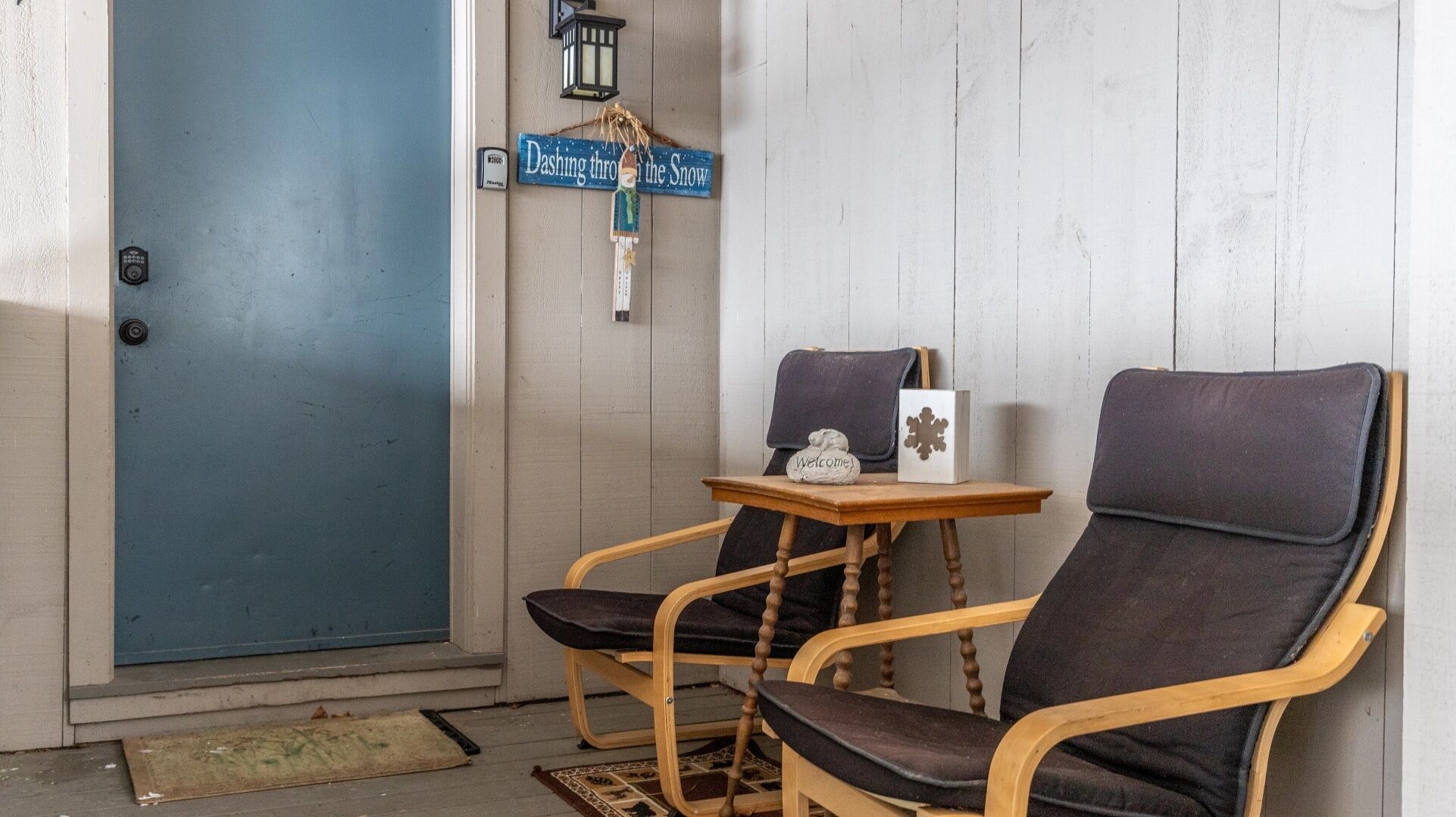 Una habitación con puerta azul, dos sillas de madera con cojines, una pequeña mesa de madera con un cartel de bienvenida y un adorno, y un farol fijado a la pared.