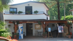 Un área de recepción con un letrero, una pérgola de madera, plantas en macetas y una mesa con sillas.