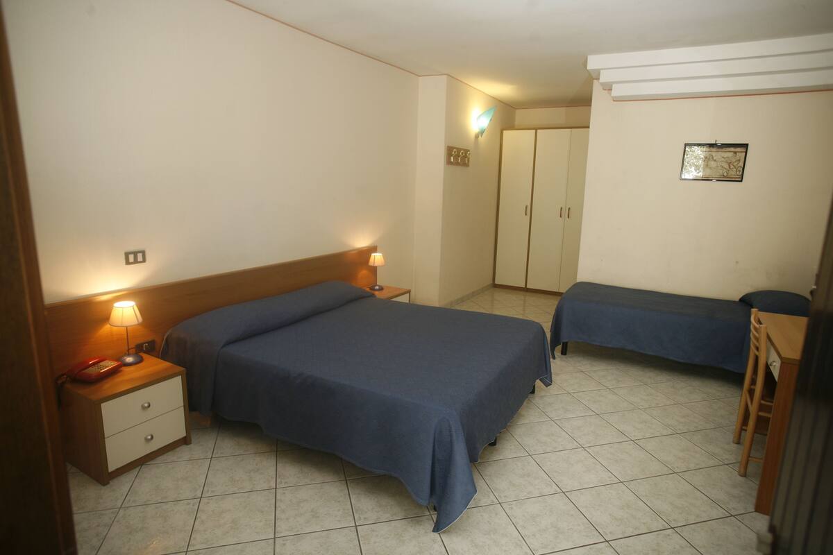 Habitación de hotel con dos camas, un escritorio, una silla, un armario y una mesita de noche con lámpara.
