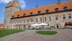 A large brick building with a red-tiled roof and a tower. There are outdoor tables and umbrellas in front of the building. - Bäckaskog Slott (Fjalkinge)