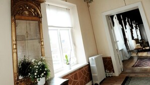 A room with a large ornate mirror, a window with white curtains, a radiator, and a wooden floor. - Bäckaskog Slott (Fjalkinge)