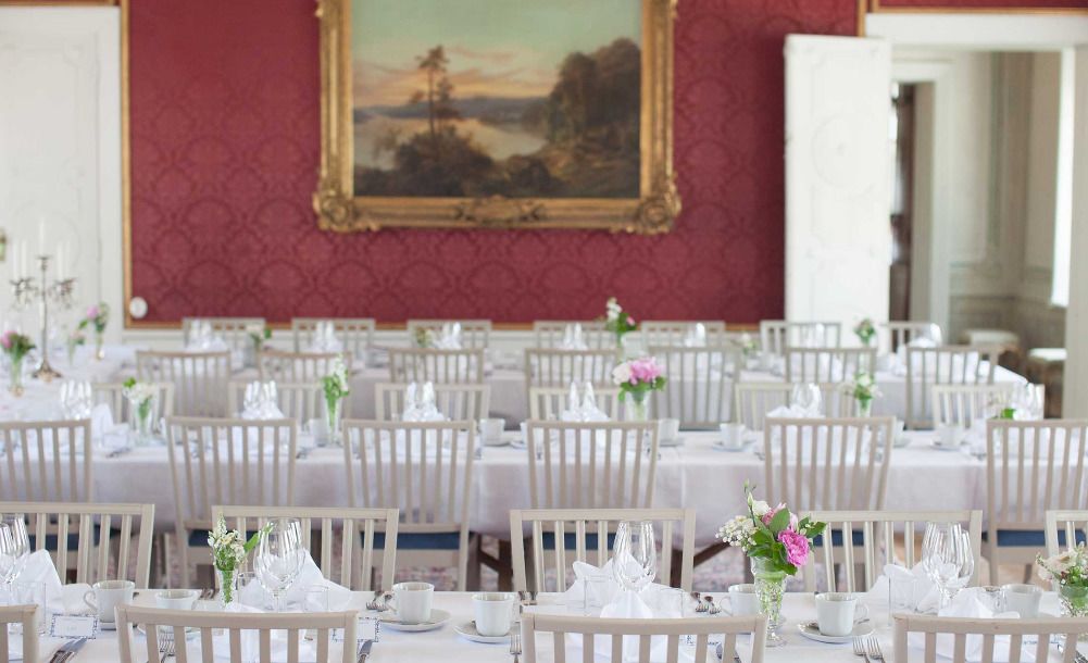 A banquet hall with white tables and chairs, a large framed landscape painting on the wall, and a red patterned wallpaper.