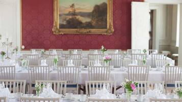 A banquet hall with white tables and chairs, a large framed landscape painting on the wall, and a red patterned wallpaper.