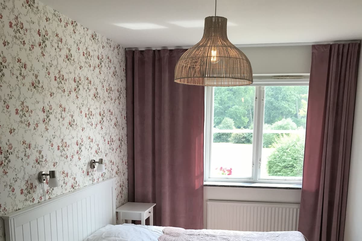 Une chambre à coucher avec un lit, une table de chevet, une lampe, une fenêtre et un papier peint à motifs floraux.