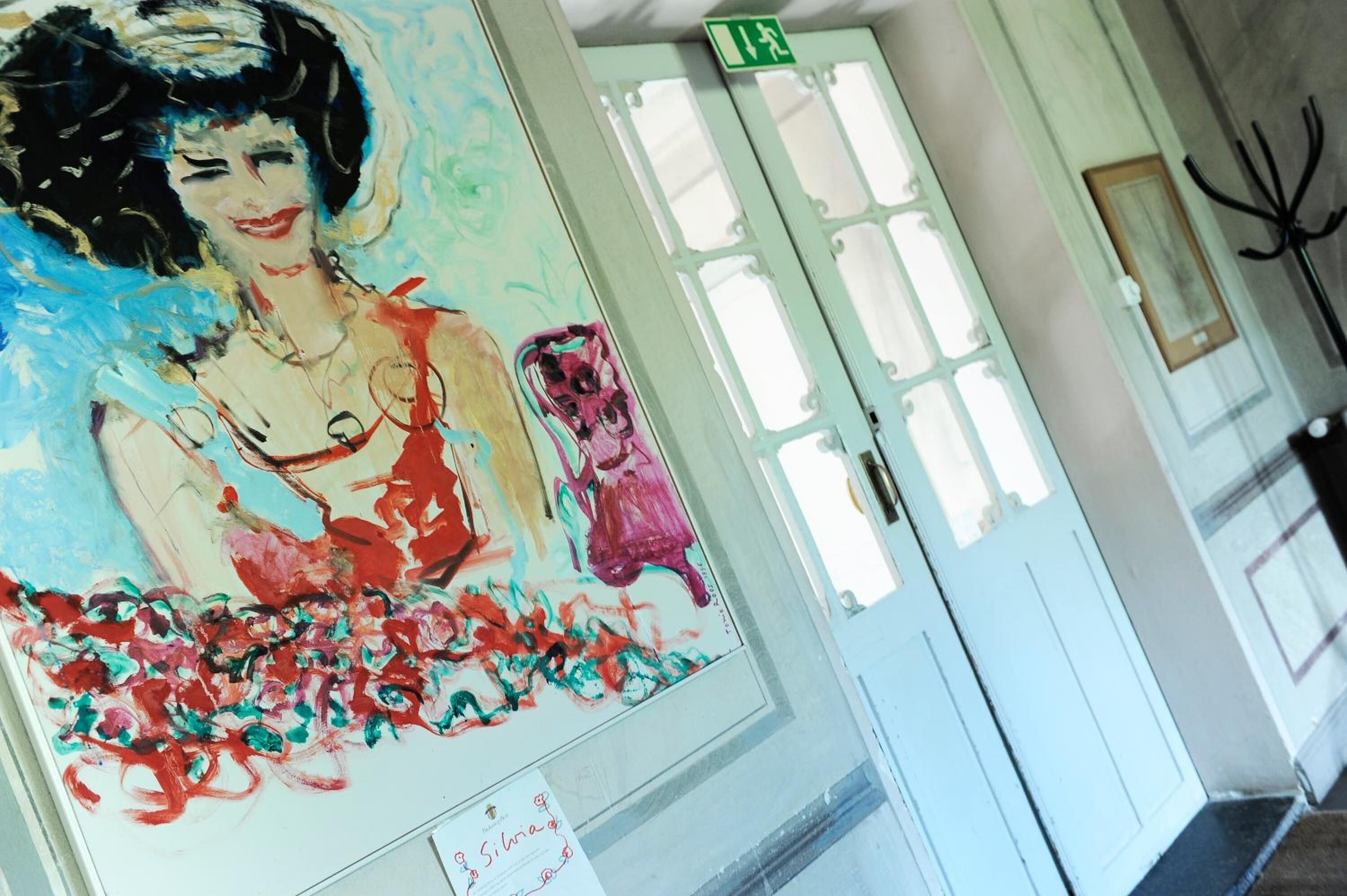 A hallway with a large abstract painting on the wall, a white door with a green exit sign, and a coat rack to the right.