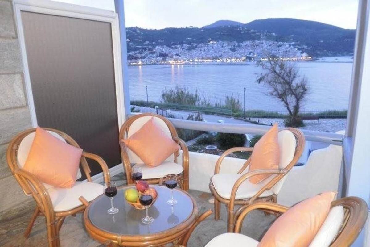 A balcony with wicker chairs and a glass table, overlooking a body of water and a distant town.