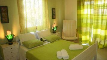 Un dormitorio ordenado con cama, almohadas, una silla y una mesita de noche con lámpara.