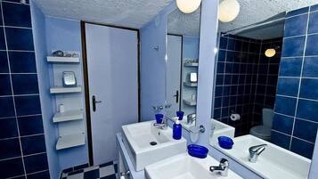 A bathroom with two sinks, a mirror, and a shower. There are shelves with toiletries and a towel rack.