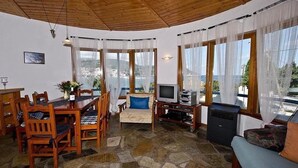 Un salon chaleureux avec un sol en pierre, des meubles en bois, un téléviseur et de grandes fenêtres donnant sur un paysage magnifique.
