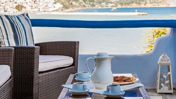 Une chaise en osier avec un coussin, une table avec une carafe, des tasses et une assiette de biscuits, le tout donnant sur une ville côtière.