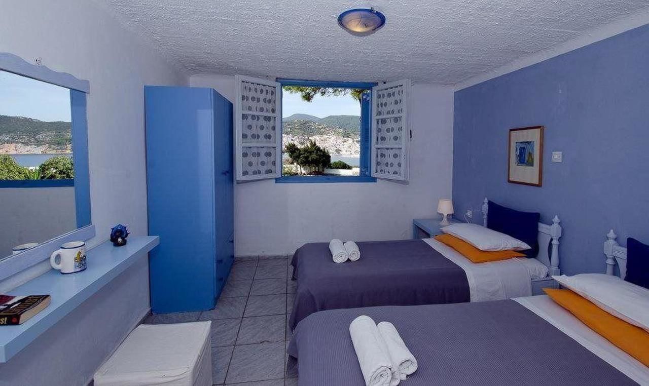 Una habitación con dos camas, una ventana con contraventanas, una mesita con una taza y un libro, y vistas al mar y a las colinas.