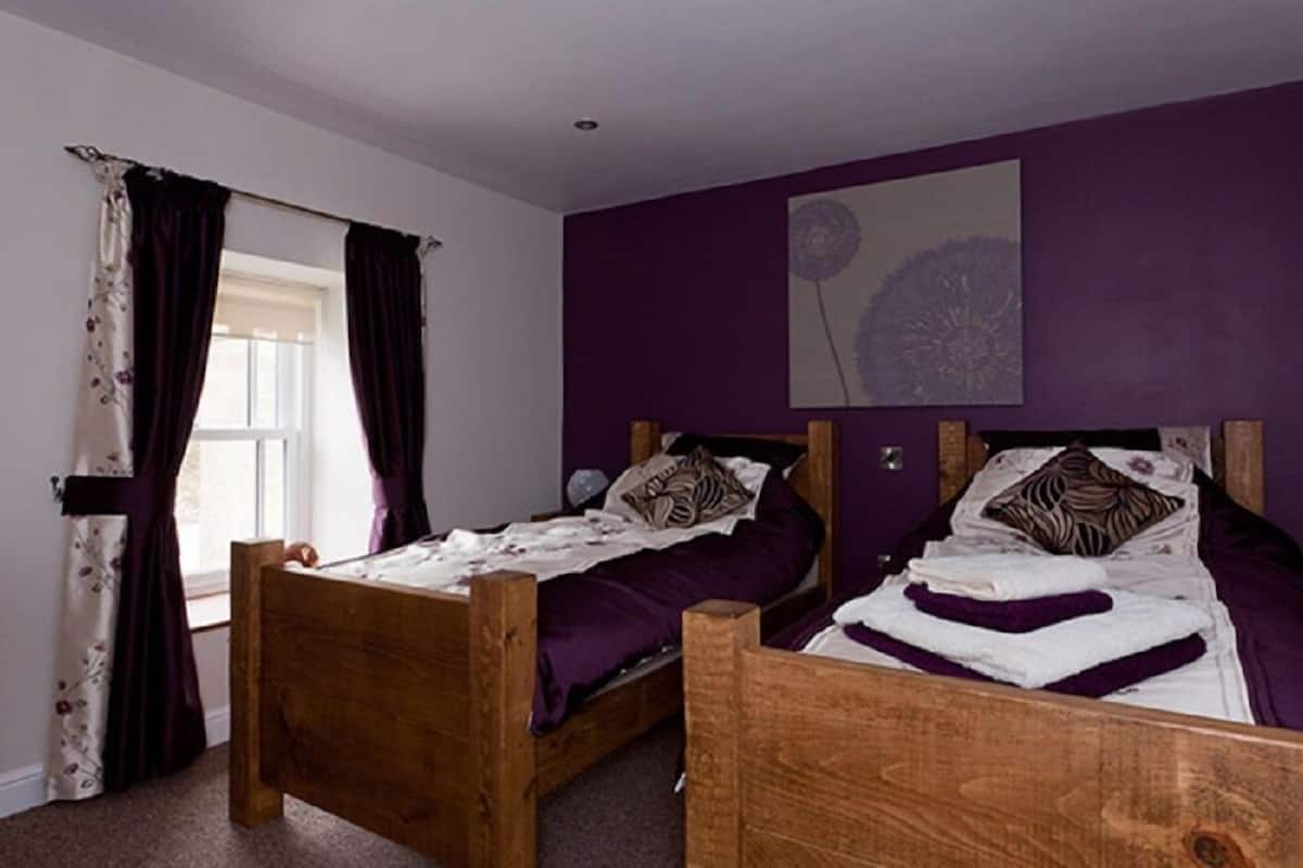 A bedroom with two wooden beds, purple bedding, and a window with curtains.