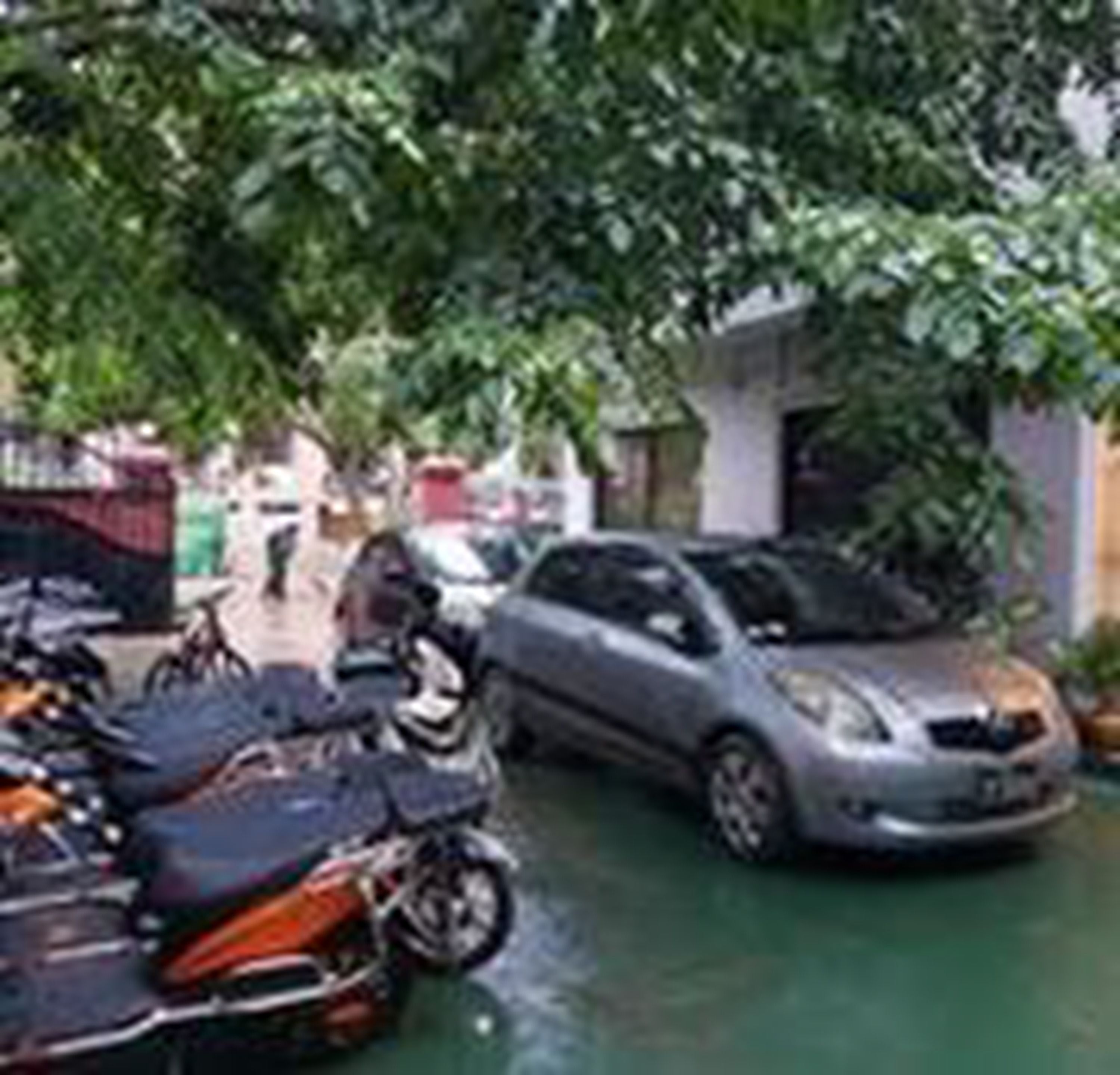 Une rue étroite, bordée de motos et d’une voiture garées, entourée de verdure et d’immeubles.