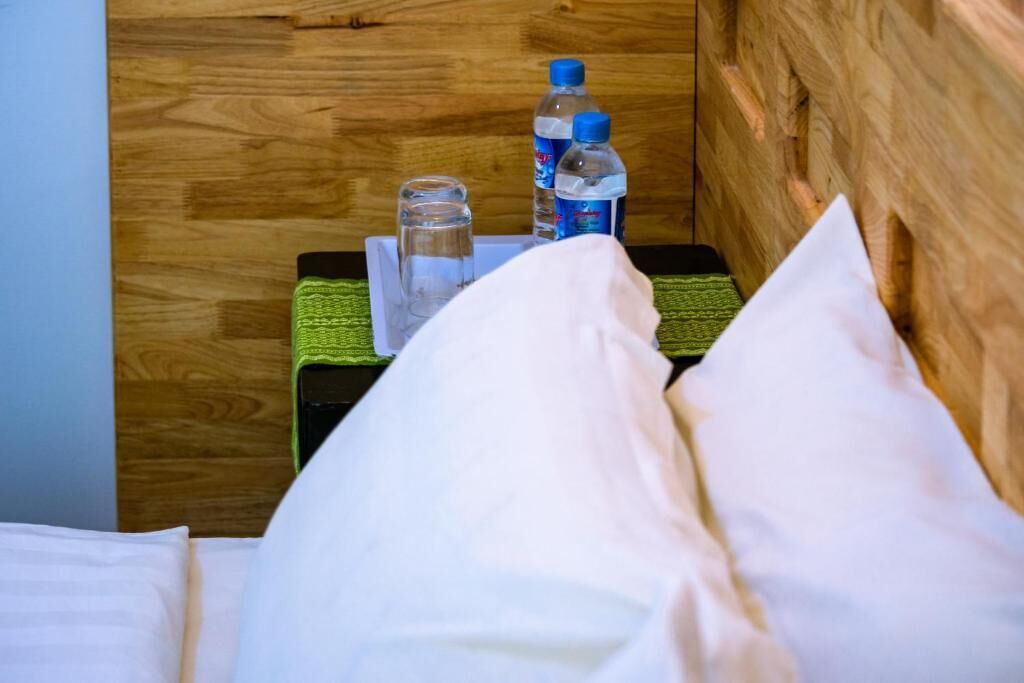 Un lit avec des oreillers blancs, un plaid vert et deux bouteilles d’eau posées sur une table de chevet en bois.