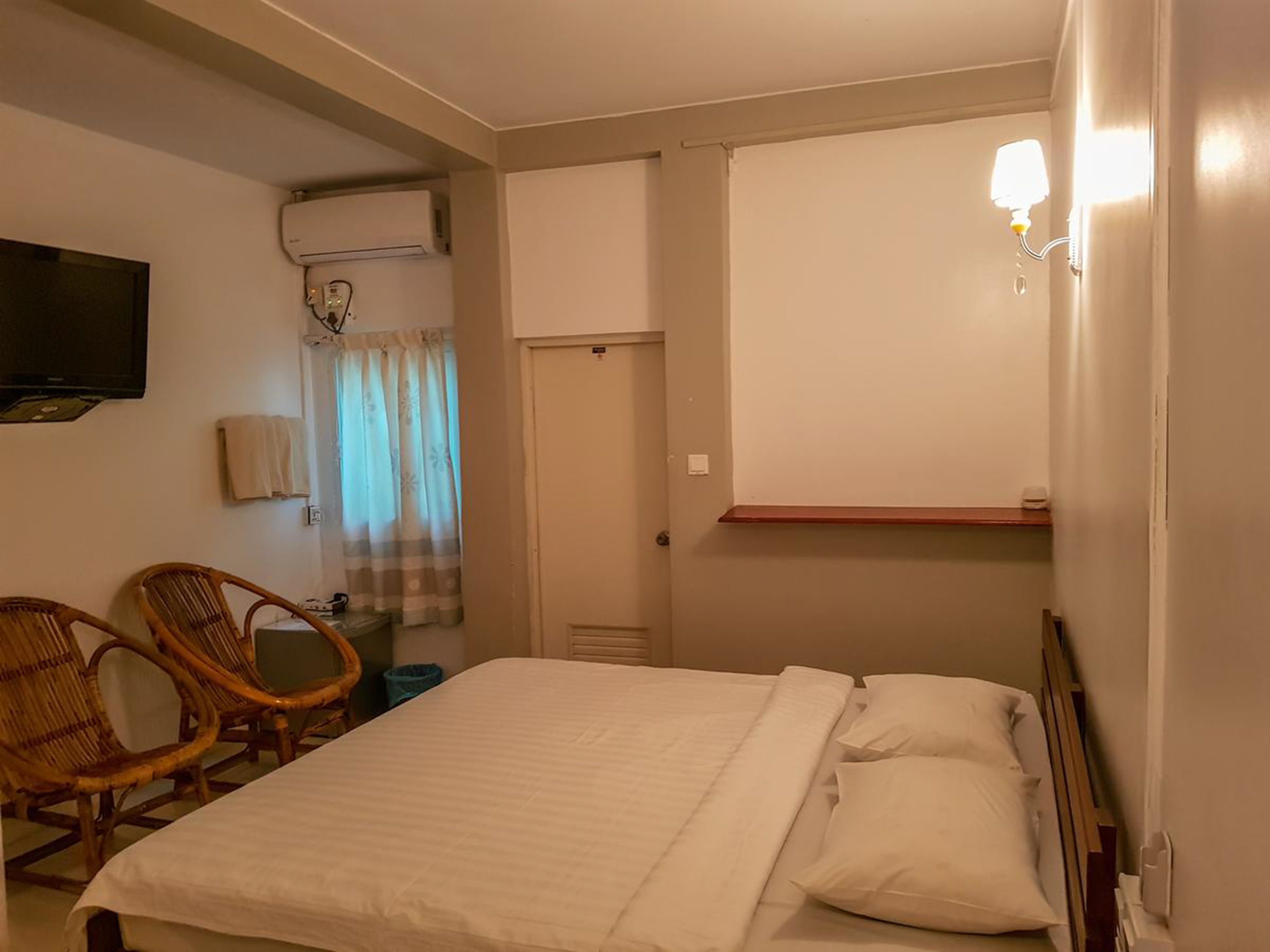 A bedroom with a bed, two wicker chairs, a television, an air conditioning unit, a window with curtains, and a bedside table.