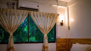 Una habitación con cama, cortinas, aire acondicionado y ventana con vistas a follaje verde.