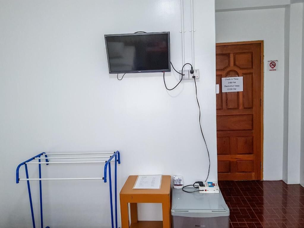 Quarto com televisão na parede, suporte de metal azul, mesa de madeira com um aviso e armário cinza.