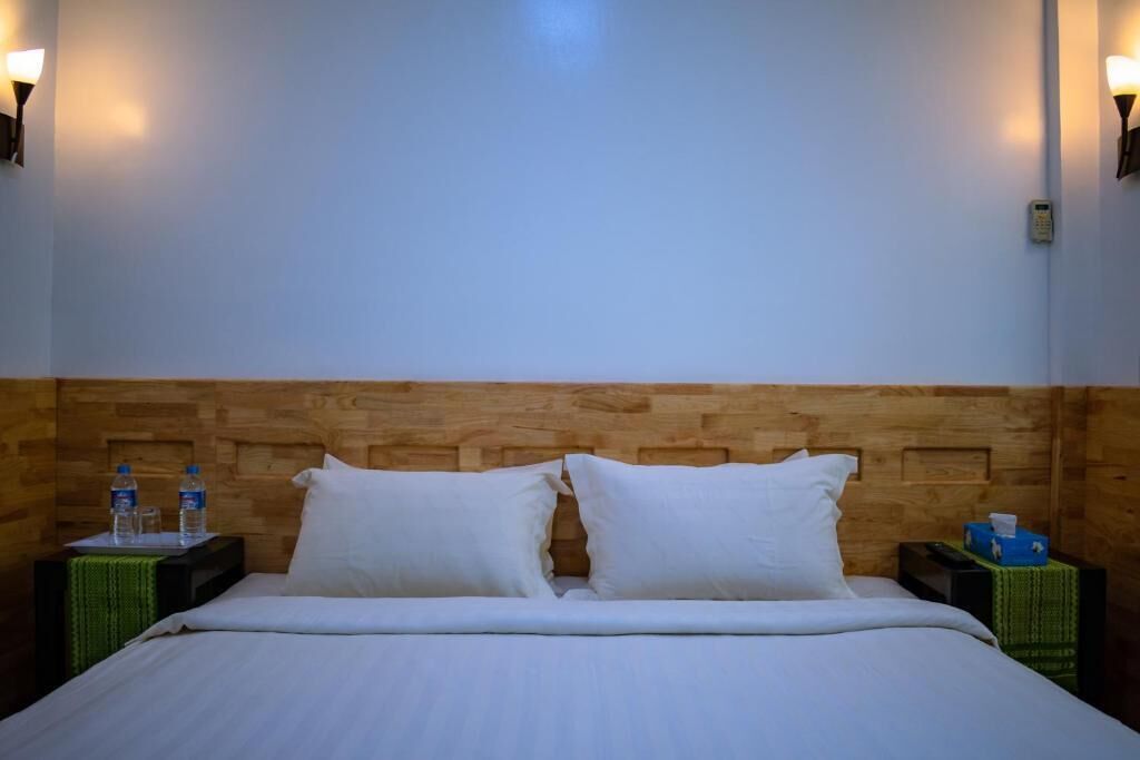 Habitación de hotel con cabecera de madera, dos almohadas blancas, cama con ropa de cama blanca, dos mesitas de noche con botellas de agua y una caja de pañuelos, y lámparas fijadas a la pared.