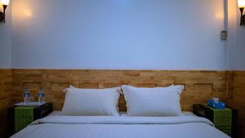 Habitación de hotel con cabecera de madera, dos almohadas blancas, cama con ropa de cama blanca, dos mesitas de noche con botellas de agua y una caja de pañuelos, y lámparas fijadas a la pared.
