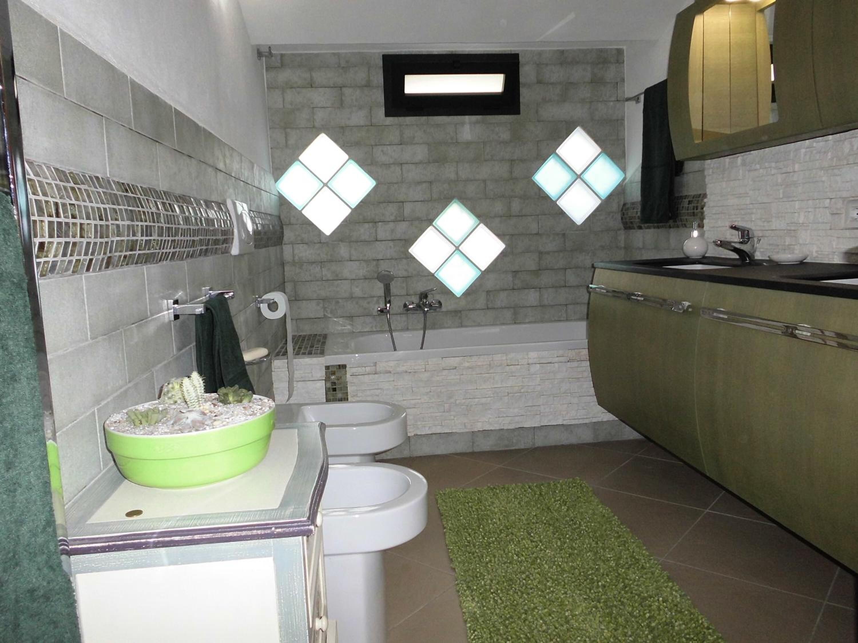 A bathroom with a toilet, bidet, and a green sink with a bowl. There is a mirror above the sink and a shower area with a glass door.