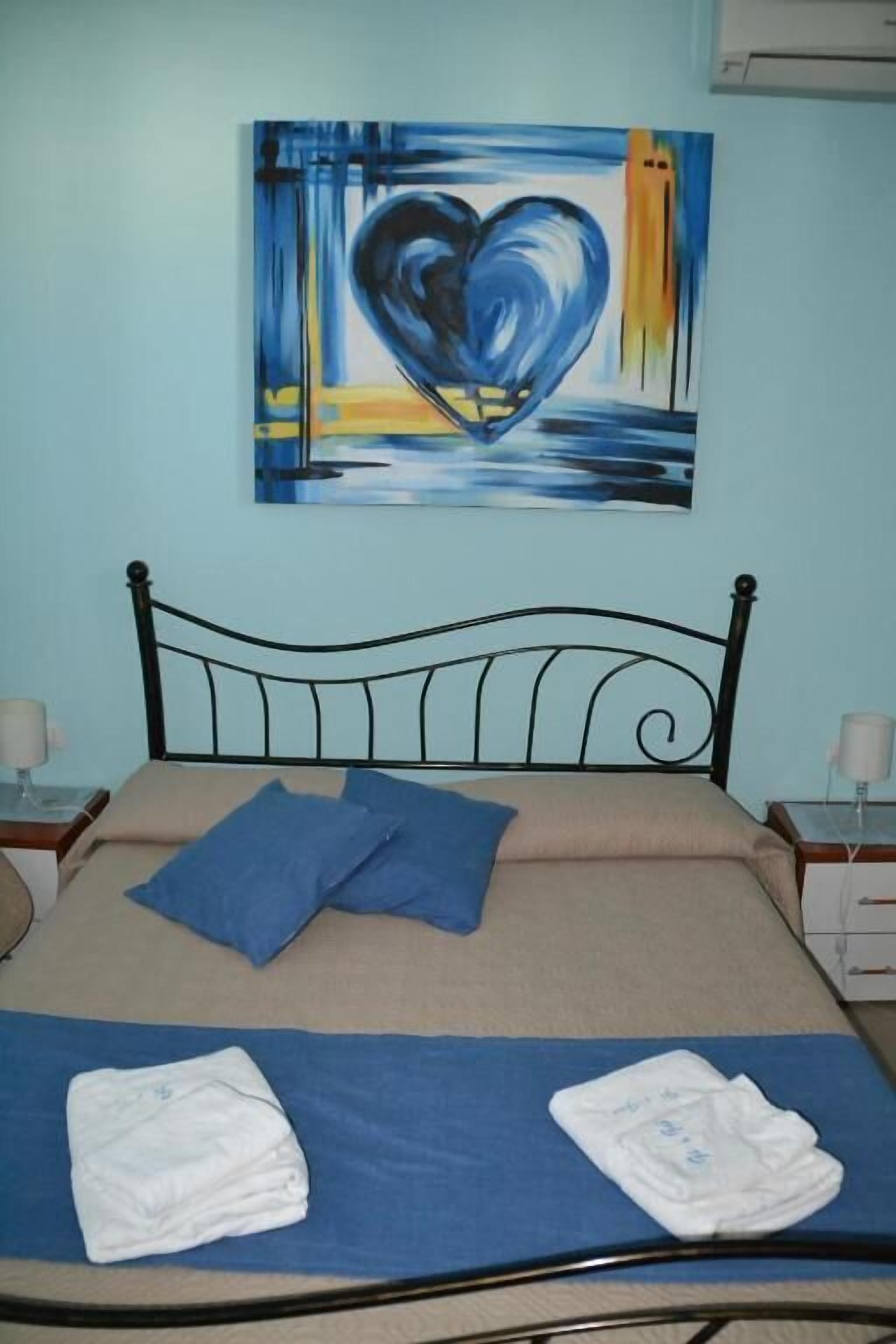Un letto con copriletto blu, due cuscini e asciugamani piegati. Sopra il letto, un quadro raffigura un cuore.