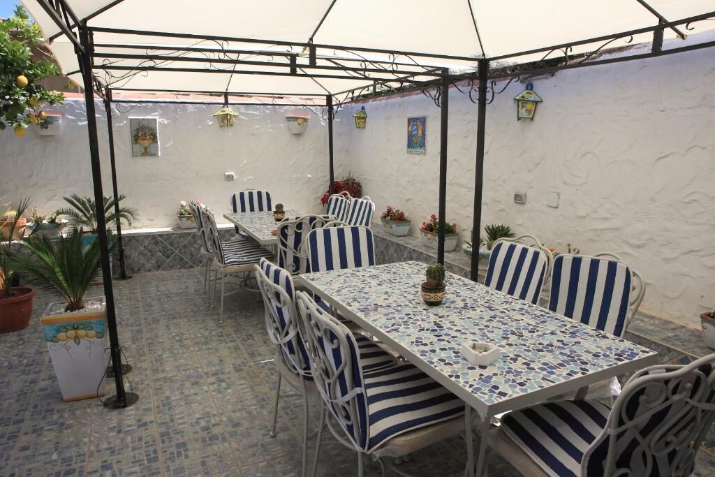 A covered patio area with a long table and striped chairs, surrounded by potted plants and a white wall with decorative elements.
