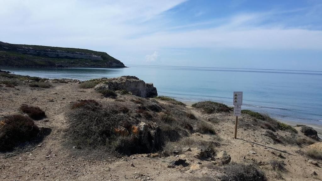 Un paysage côtier avec un sentier de terre menant à la mer, un affleurement rocheux et un panneau indicateur.