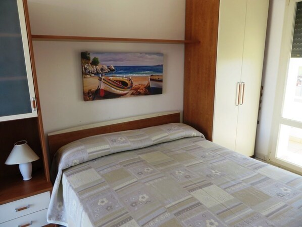 A bedroom with a bed, a painting of a beach scene, a nightstand, and a lamp.