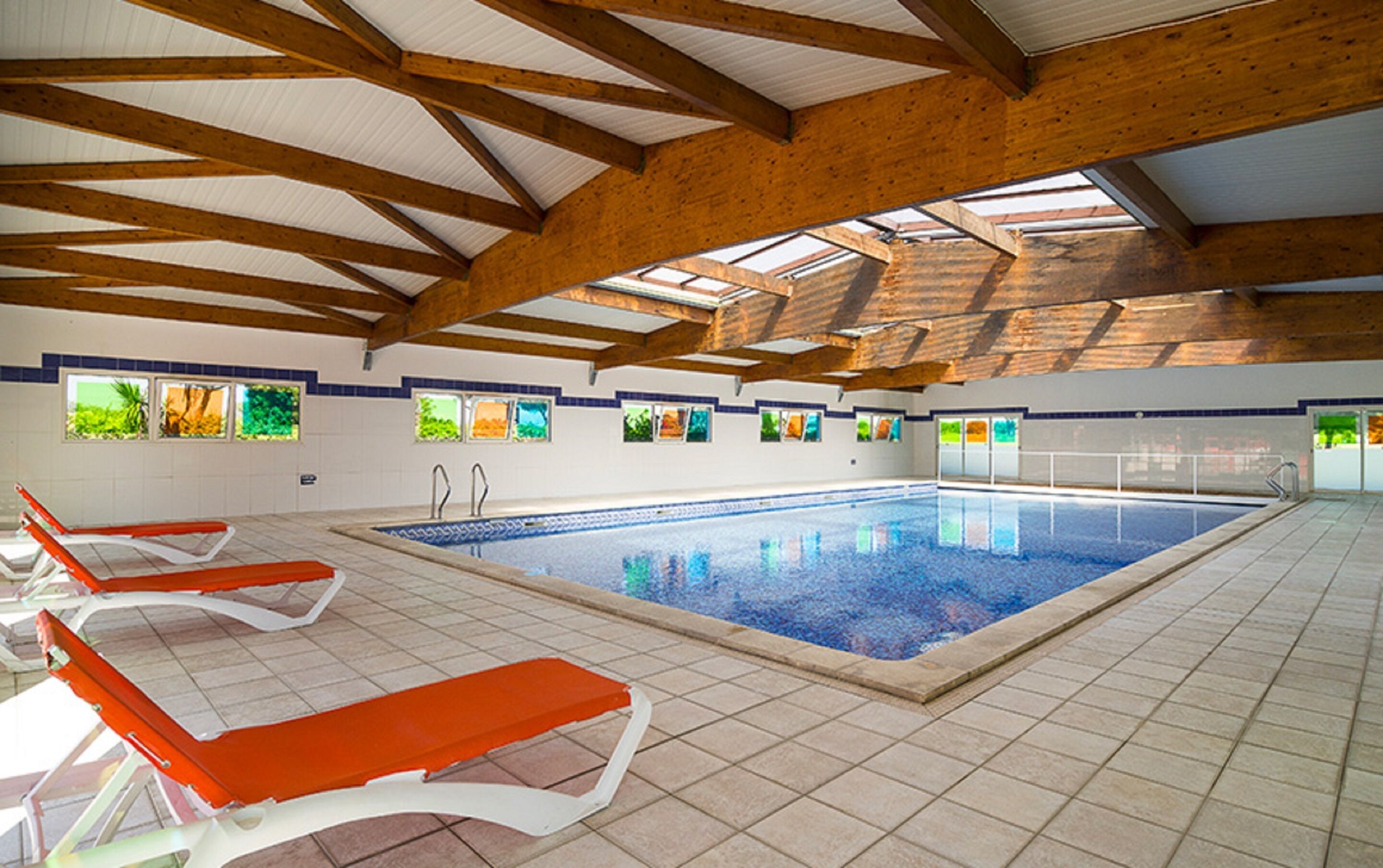 An indoor swimming pool with tiled flooring, wooden beams, and multiple windows with colorful views.