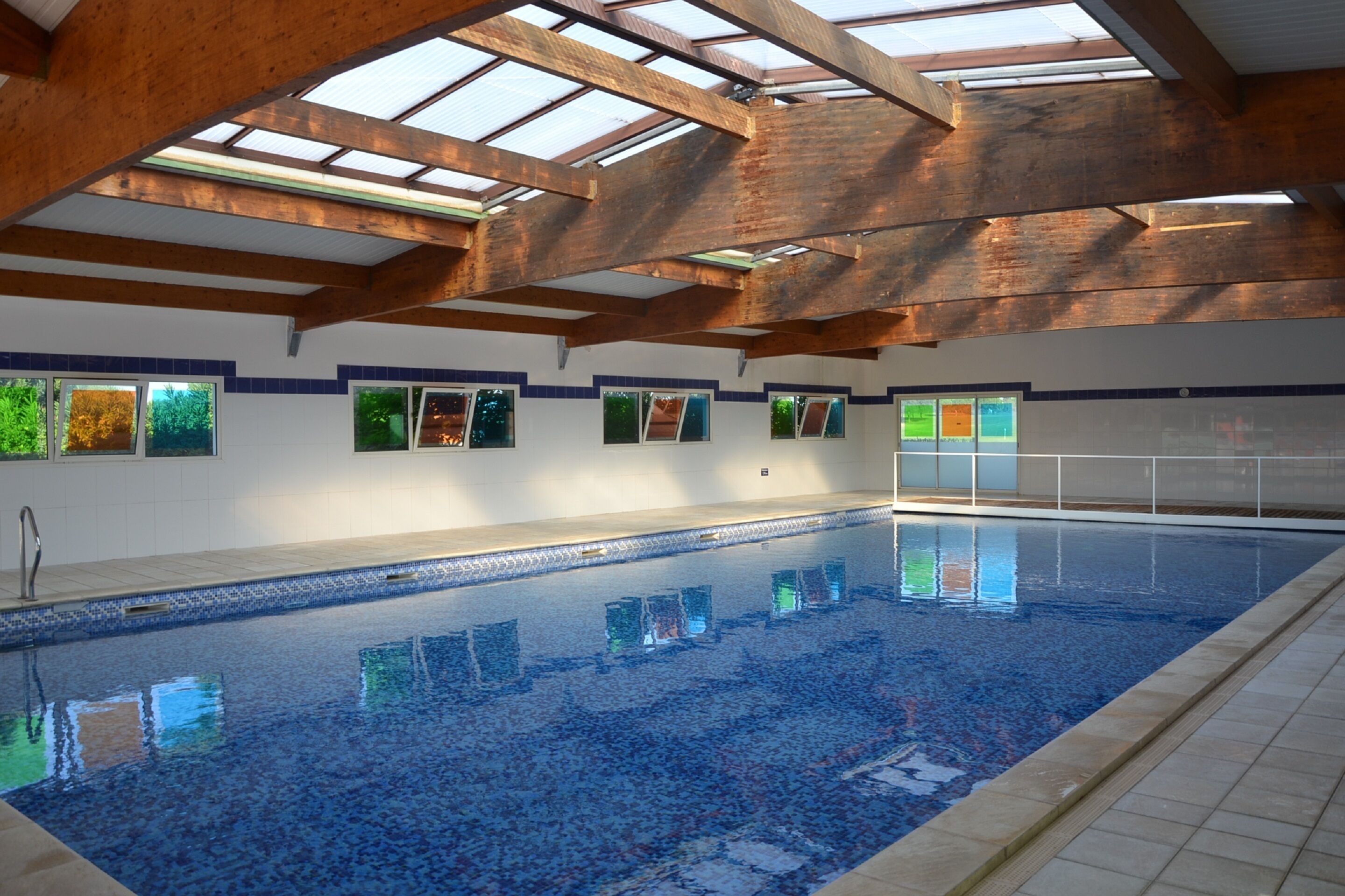 An indoor swimming pool with a tiled floor, wooden beams, and windows with green foliage views.