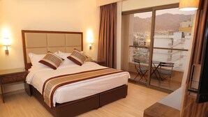 A hotel room with a large bed, a bedside table, a chair, a TV, and a balcony with a view of buildings and mountains. - Lacosta Hotel (Aqaba)