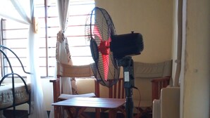 A room with a standing fan, a wooden table, chairs, and a window with curtains.