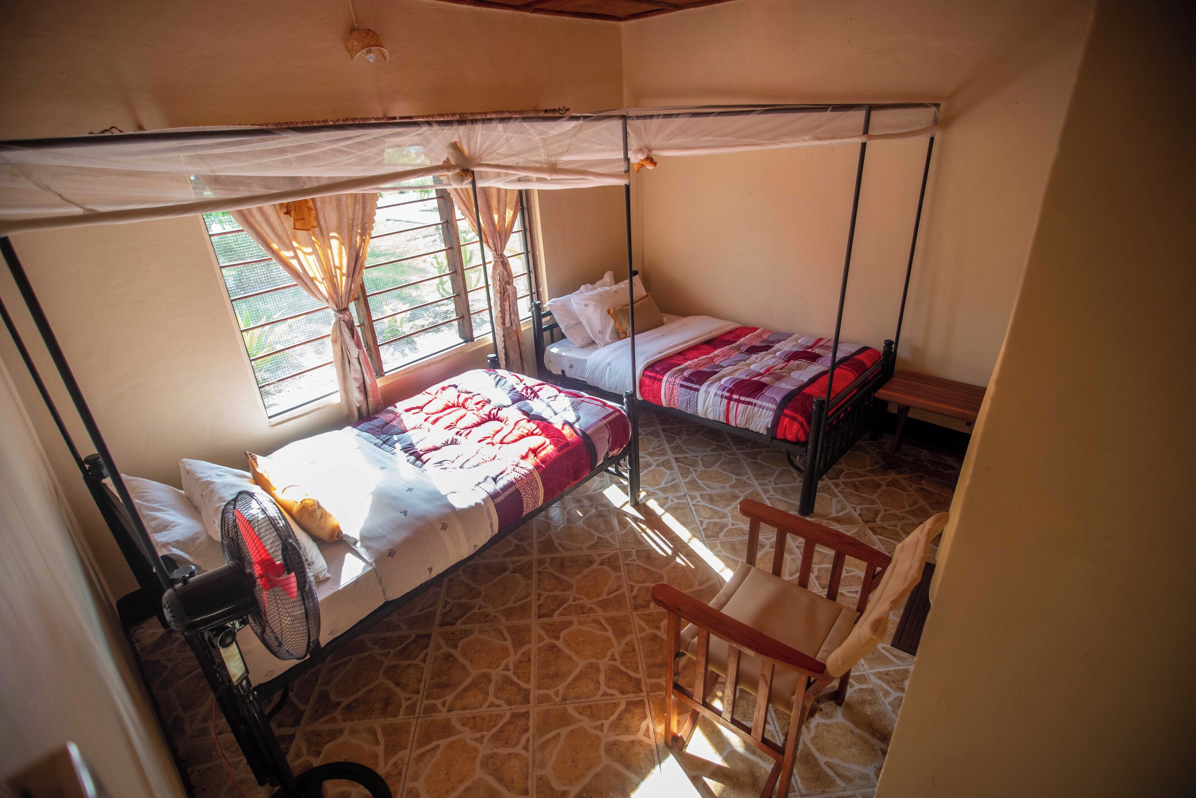 Una habitación con dos camas individuales, una silla, un ventilador de pie y una ventana con cortinas.