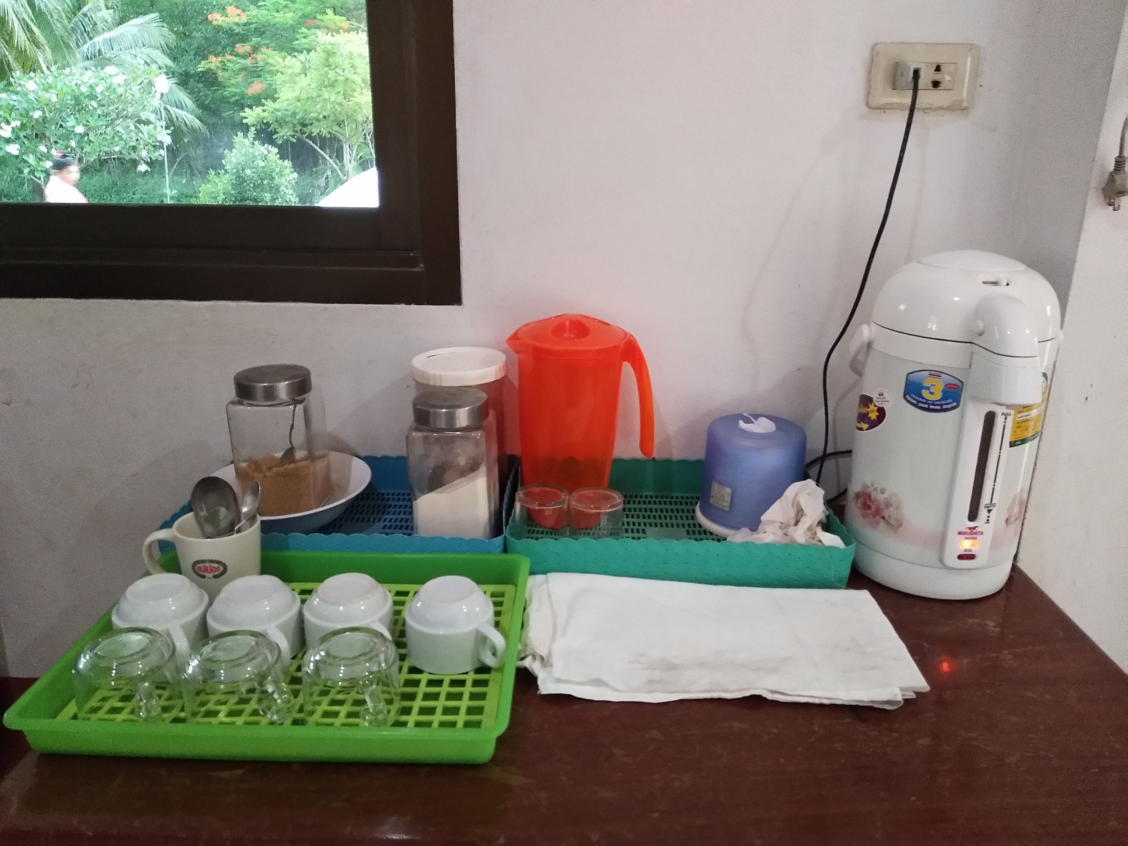 Una encimera de cocina con diversos utensilios, una ventana con vistas a la vegetación y un enchufe.