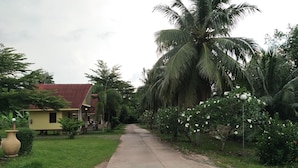 A rural road leading to a yellow house with a red roof, surrounded by lush greenery and palm trees. - Happy Home Resort (Khlong Yai)
