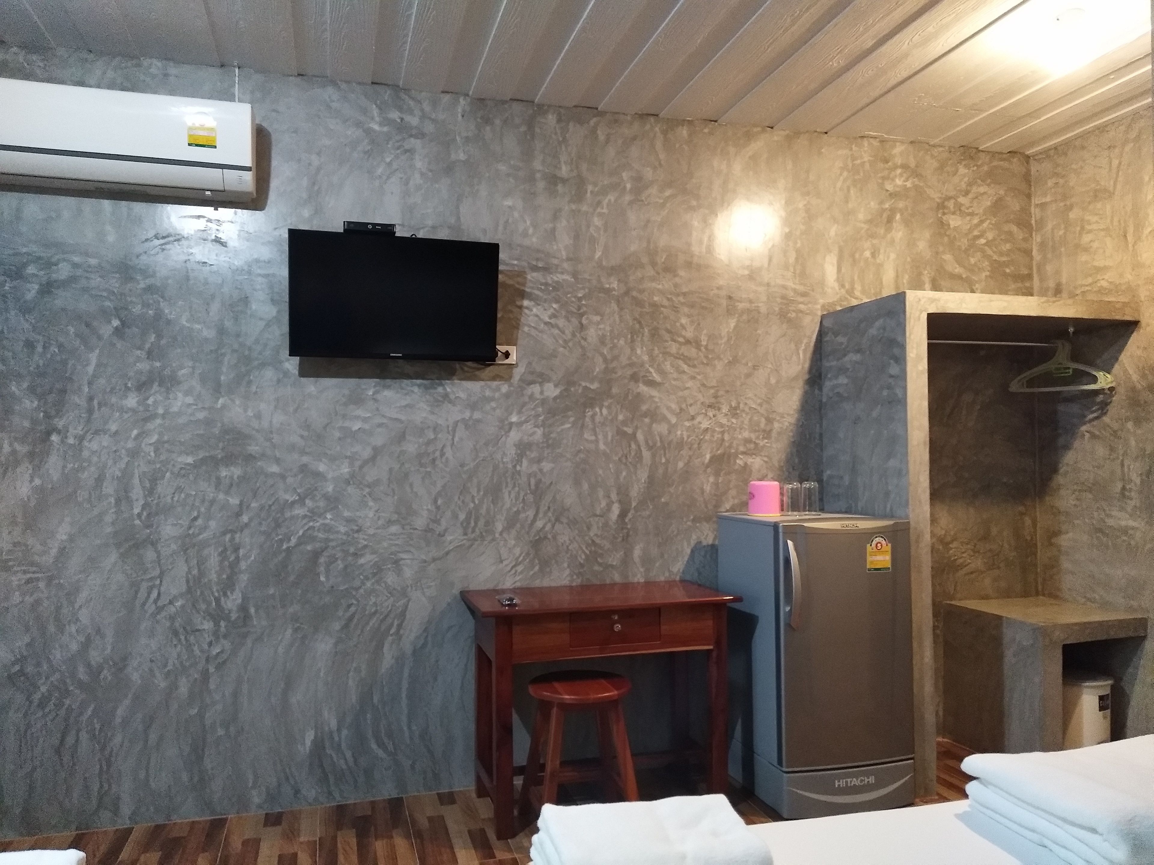 Une pièce de taille modeste avec un mur décoré d’un motif imitant le marbre, un téléviseur à écran plat fixé au-dessus d’un bureau en bois, un réfrigérateur Hitachi et un petit tabouret.