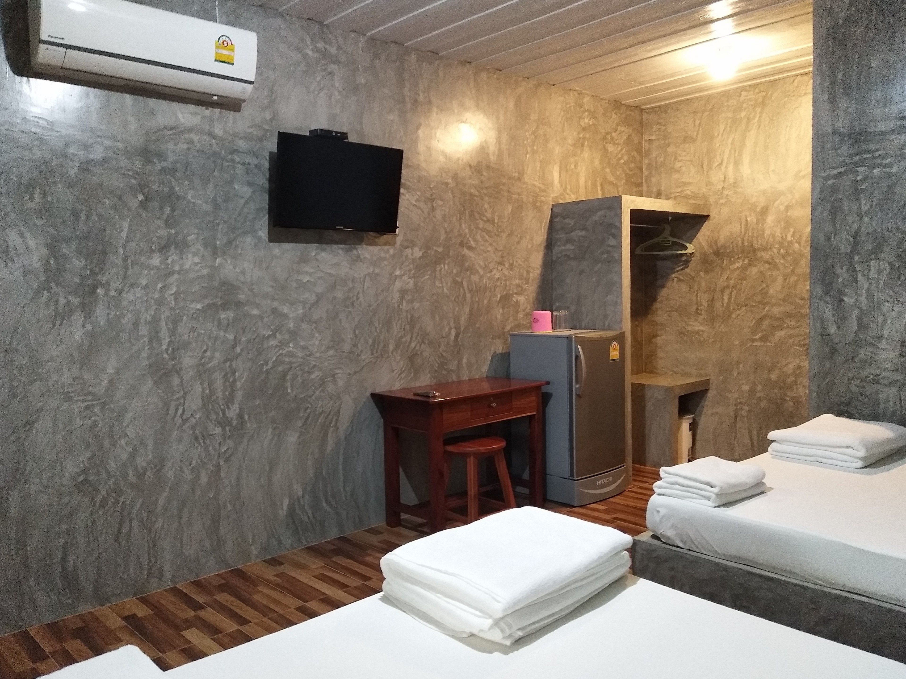 A compact hotel room with a wooden floor, a bed with white linens, a desk with a chair, a small refrigerator, and a television mounted on the wall.