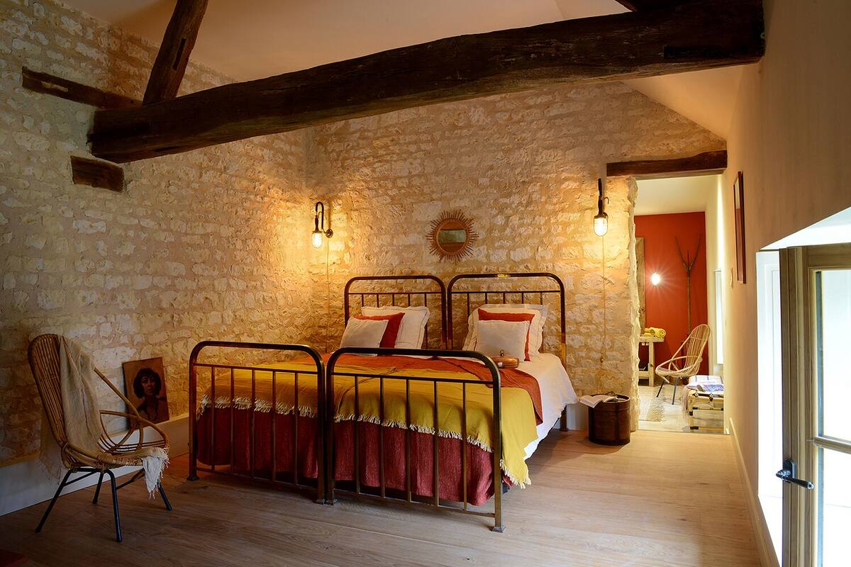Un dormitorio rústico con pared de piedra, techo de madera, estructura de cama de metal y una silla de mimbre.