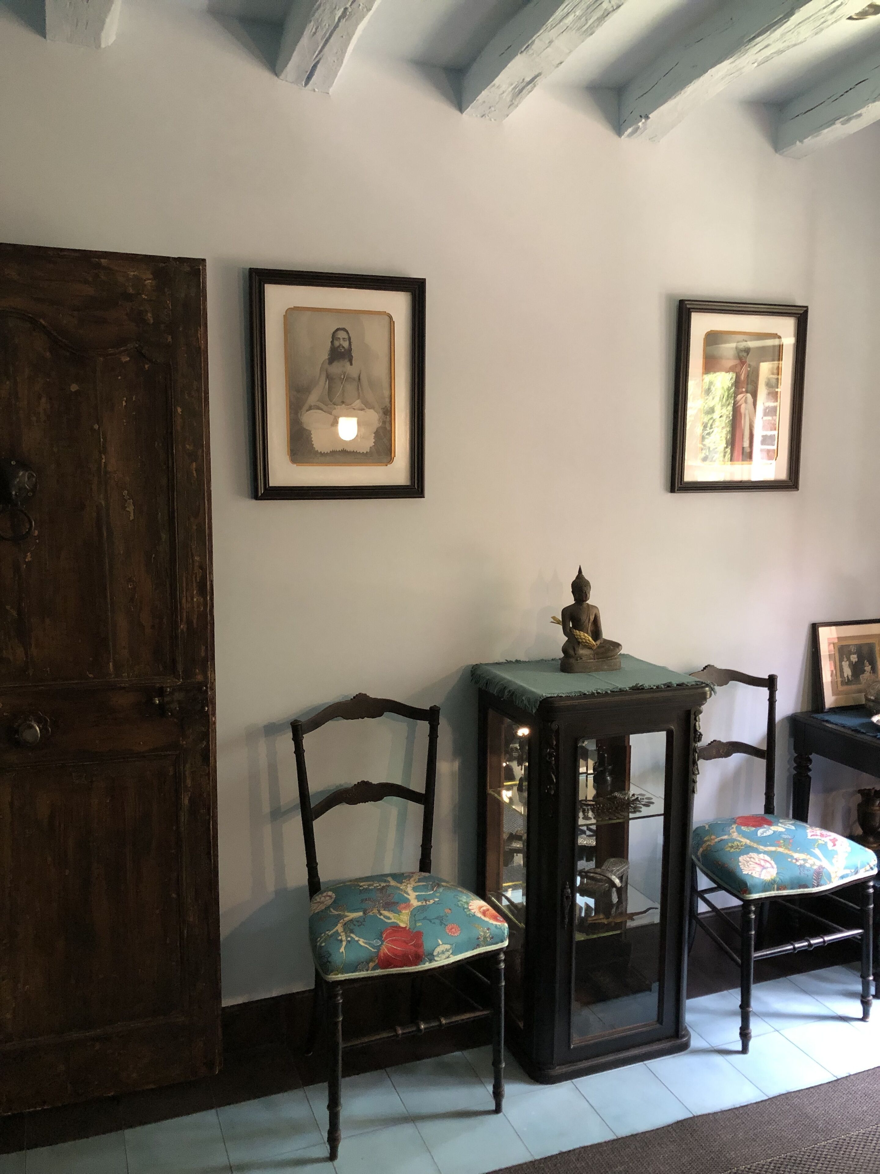 Una habitación con puerta de madera, dos cuadros en la pared, una mesita con una estatua de Buda y dos sillas con tapicería floral.