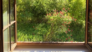 A room with a large window, a framed artwork above it, and a view of a garden with red flowers. - Domaine de l'Ocrerie (Pourrain)
