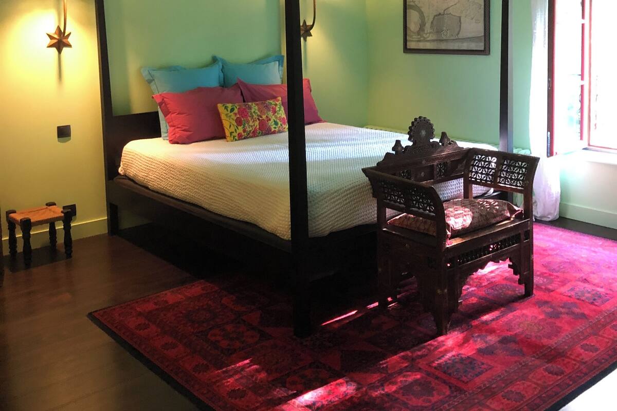 Una cama con dosel, un tapete estampado en rojo y negro, una silla de madera y una mesita.