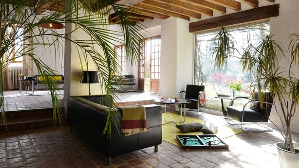 A spacious living room with a black sofa, a coffee table, and a dining area. There are large windows allowing natural light and a view of the outdoors. - Domaine de l'Ocrerie (Pourrain)